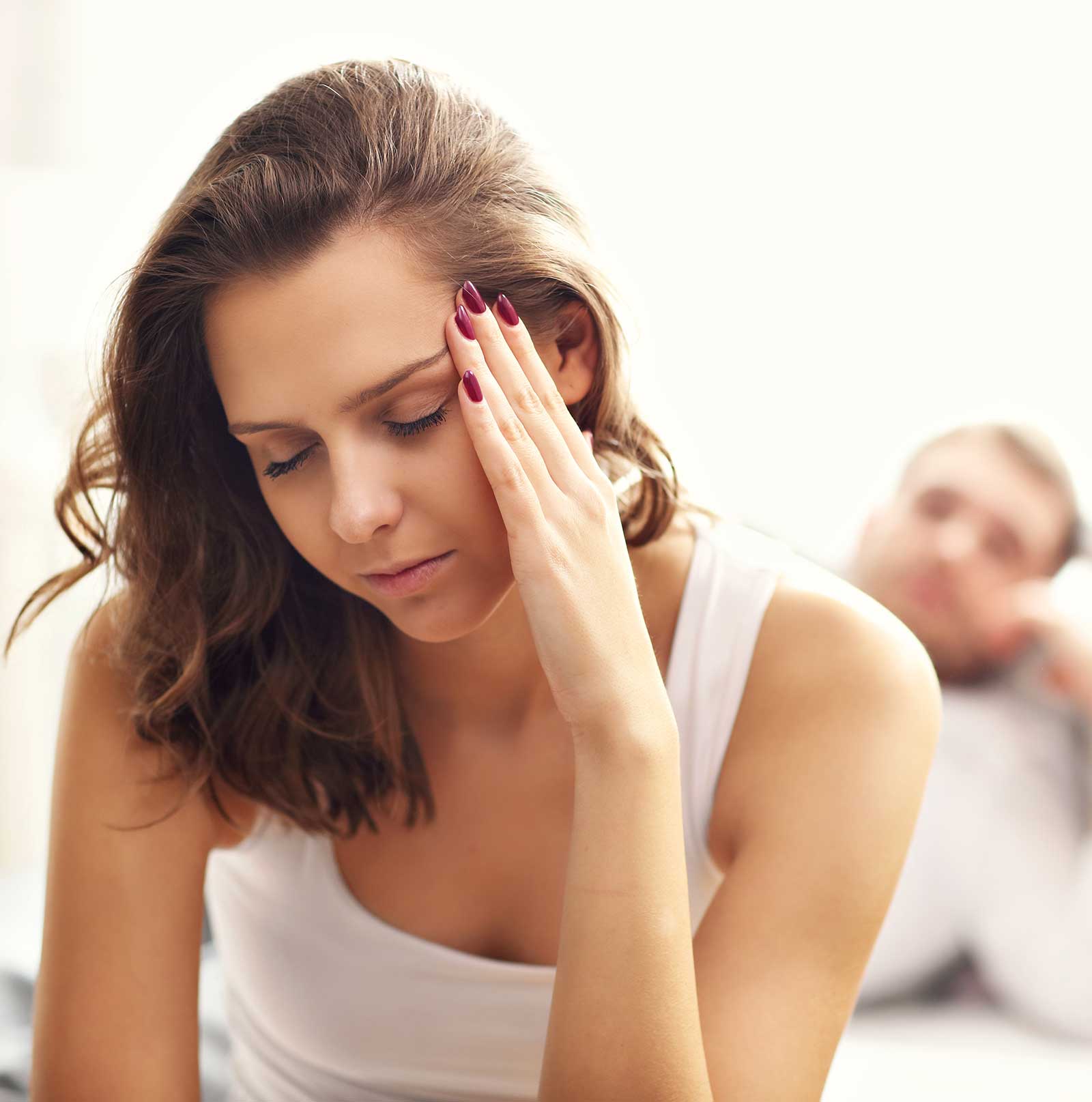 A photo of a woman holding her left temple in apparent pain. Behind her, out of focus a man looks at her. The photo represents Vaginal Dryness & Painful Intercourse, one of the conditions treated with Vaginal Rejuvenation at The Women's Clinic