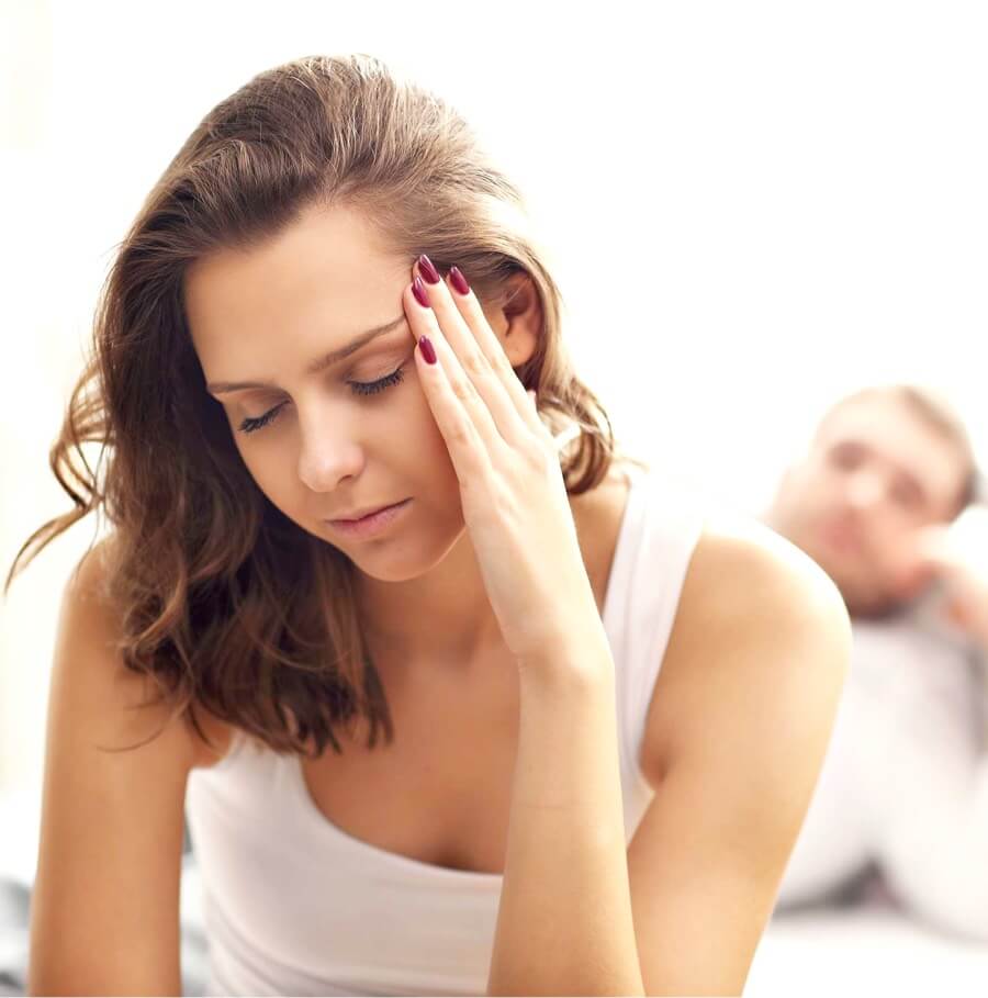 A photo of a woman holding her left temple in apparent pain. Behind her, out of focus a man looks at her. The photo represents Vaginal Dryness & Painful Intercourse, one of the conditions treated with Vaginal Rejuvenation at The Women's Clinic