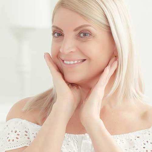 A photo of a woman smiling as she looks at herself in the mirror. This page is for lines & wrinkles, loose, ageing & sun-damaged skin on the face, neck & décolleté, some of the skin conditions treated at The Women's Clinic